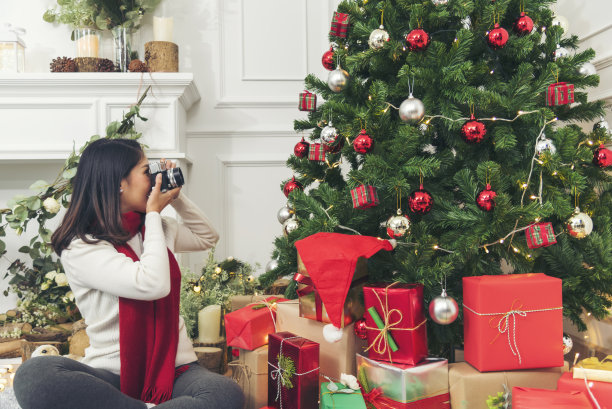 女摄影师拍照片圣诞节装饰小玩意儿球圣诞节树在节日冬季庆祝。亚洲妇女用相机拍照圣诞树圣诞礼物礼盒在舒适的客厅图片下载