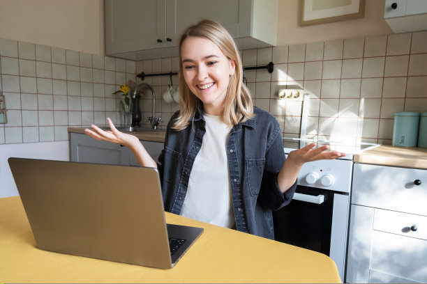 一个年轻的金发女孩微笑着坐在厨房里的笔记本电脑旁，在家办公。远程工作的概念，家庭主妇的生活图片下载