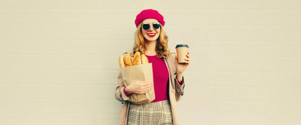 画像快乐微笑的年轻女子拿着杂货店购物纸袋长白面包法面包和杯咖啡在灰色背景，空白的复制空间为广告文字图片下载