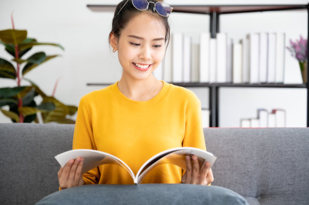 快乐的年轻亚洲女人在家里客厅的沙发上看书图片下载