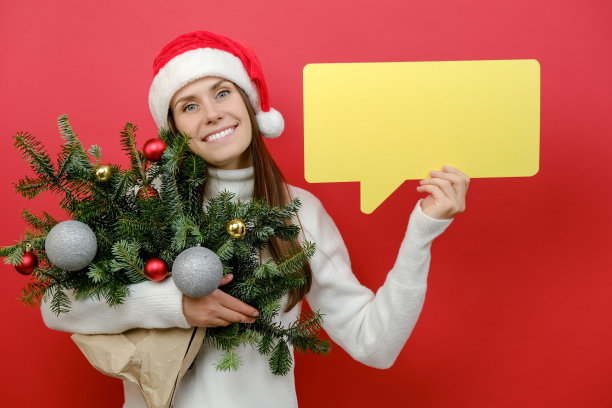画像快乐的年轻女性在圣诞帽拿着一束云杉树枝和黄色空空白说云，发言气泡摆上红色的背景。新年喜庆喜庆佳节概念图片下载