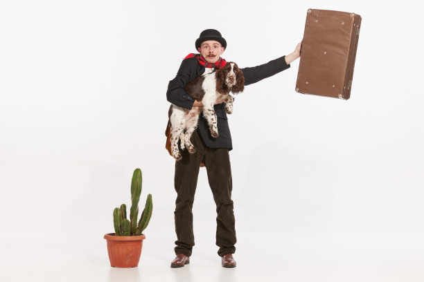 肖像时尚的年轻人在外套和帽子，抱着狗和举起手与手提箱孤立在白色背景图片下载