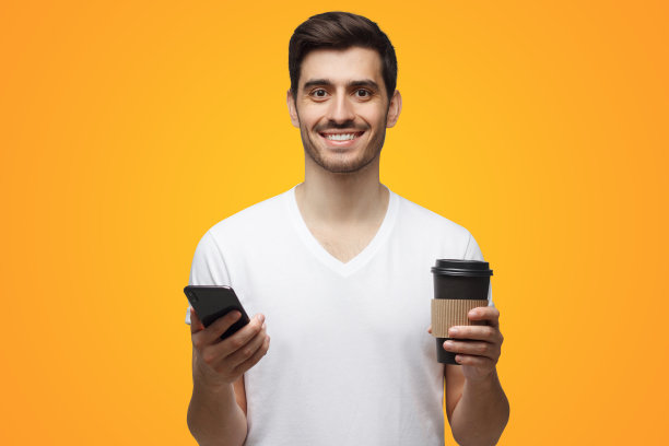 一名男子看着相机，一只手拿着手机，另一只手拿着咖啡杯，背景是黄色图片下载