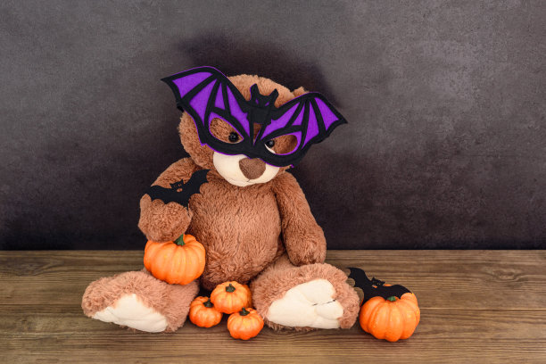 有趣的棕色泰迪熊与蝙蝠形状的面具和南瓜在旧木桌黑墙图片下载