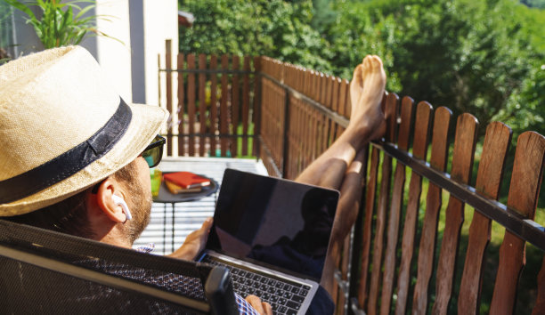 戴着帽子和太阳镜的男性在阳台上用耳机听音乐，用笔记本电脑。图片下载