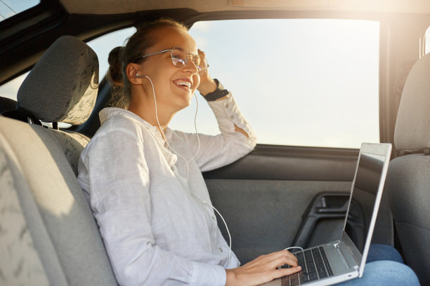 侧面肖像的吸引力开朗快乐的妇女坐在汽车后座和笔记本电脑工作，听着耳机音乐，看向别处，笑得很开心，享受她的工作在车辆。图片下载