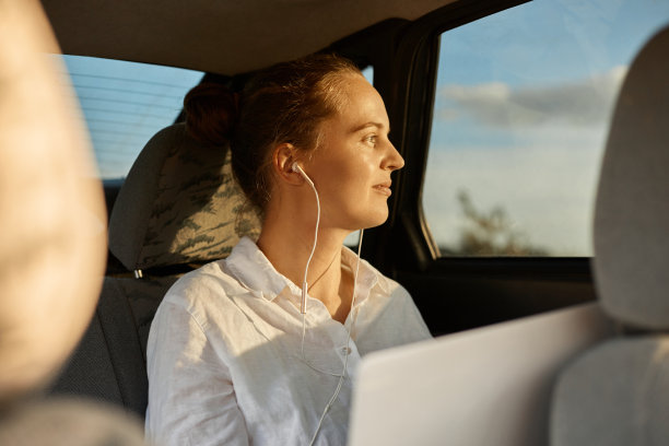 侧面肖像美丽的女性自由职业者穿着白衬衫坐在汽车后座上，用笔记本电脑工作，听音乐，看着车窗。图片下载