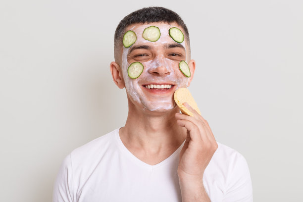 穿着白色t恤的英俊男子，戴着面膜、黄瓜片和海绵，在灰色背景下，满意地看着镜头。图片下载