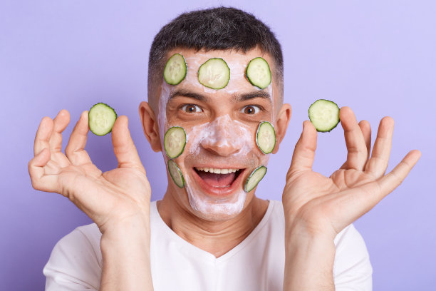 照片中震惊的年轻男子穿着白色t恤，脸上敷着黄瓜片的面膜，手里拿着蔬菜，正在做皮肤护理，被紫色的背景隔离开来。图片下载