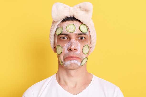 悲伤不安的男子穿着白色t恤和带蝴蝶结的发带，感到悲伤，撅着嘴看着镜头，戴着口罩和黄瓜在黄色背景下摆姿势。图片下载