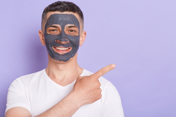 室内拍摄的男子在白色t恤和化妆面具在他的脸上，做抗衰老的程序，指着旁边的蚂蚁复制空间的广告或促销文字。图片下载