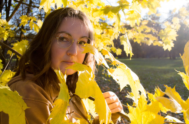 在秋日阳光明媚的日子里，一个戴着圆眼镜的美丽年轻女子的肖像在黄色的枫叶丛中图片下载