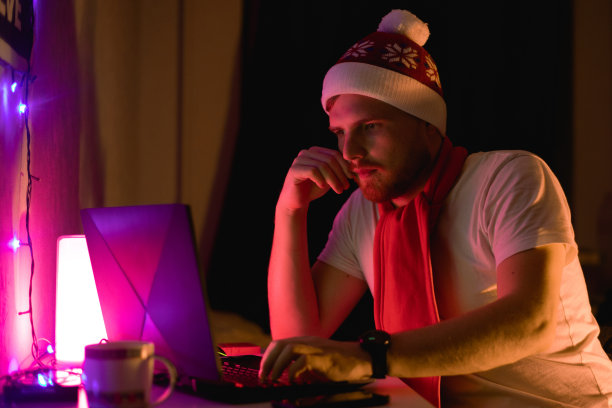 新年男性画像快乐的大胡子人戴着红色圣诞老人帽谁使用笔记本电脑工作。圣诞节庆祝的概念。室内摄影，男人带着帽子和电脑图片下载