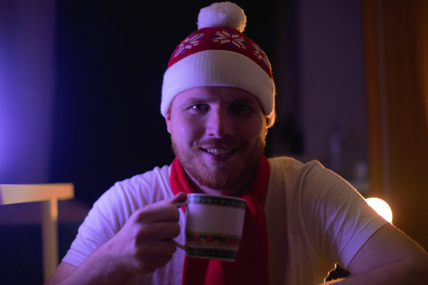 人在室内戴着圣诞红的圣诞老人帽喝着热腾腾的新年饮料，从杯子里倒出茶或咖啡，家中气氛在霓虹灯下。男人肖像摄影图片下载