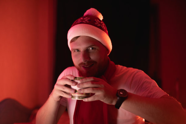 人在室内戴着圣诞红的圣诞老人帽喝着热腾腾的新年饮料，从杯子里倒出茶或咖啡，家中气氛在霓虹灯下。男人肖像摄影图片下载