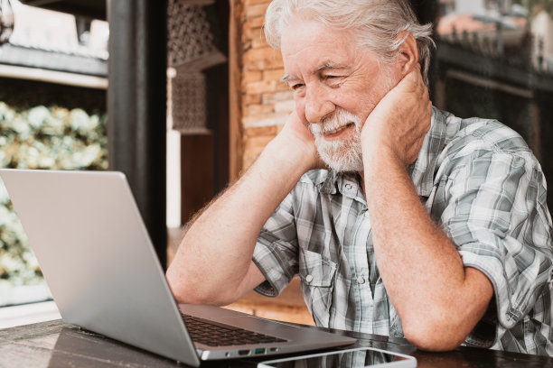 大胡子老人坐在户外的木桌上用电脑阅读电子邮件。英俊的老年男性在笔记本电脑上浏览互联网图片下载