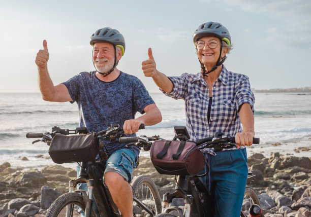 快乐的老年白人夫妇骑在卵石海滩上的电动自行车手势积极的信号。真实的老年退休生活理念图片下载