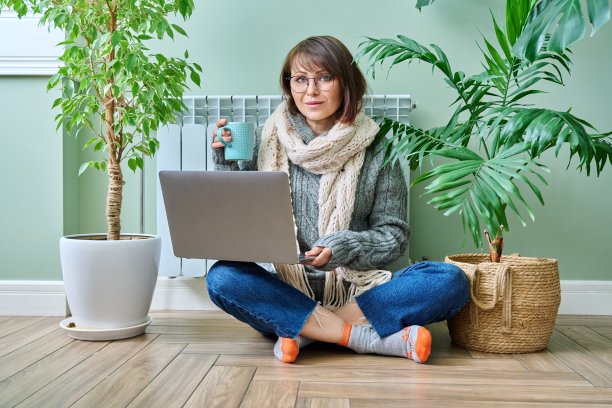 穿着暖和衣服的妇女坐在暖气散热器附近，使用笔记本电脑图片下载