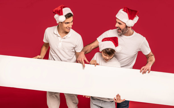 爸爸和两个儿子拿着一块白色的空板子，在红色的画室背景上摆姿势，戴着圣诞老人的帽子图片下载