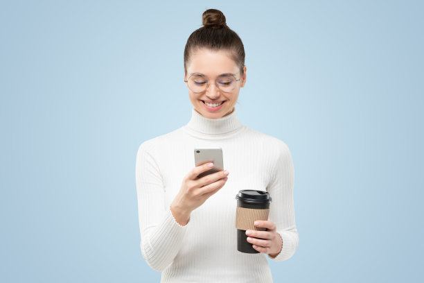 年轻迷人的女人戴眼镜和白色高领毛衣，拿着电话和咖啡杯图片下载