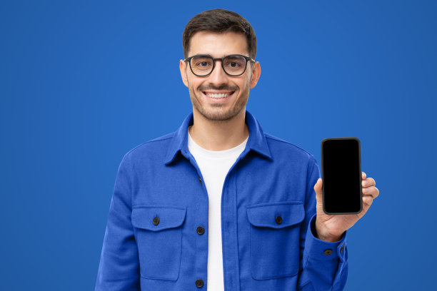 年轻的微笑的男人在休闲衬衫显示空白屏幕的智能手机在手中，孤立的蓝色图片下载