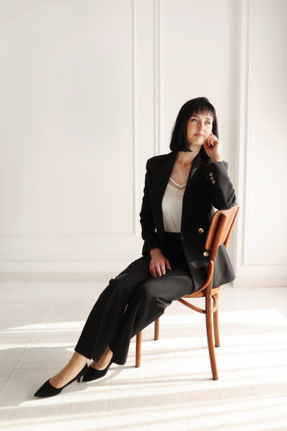 迷人的商业女性坐在木椅上，看着镜头。黑发短发的女士穿着黑色办公套装和白色衬衫，白色墙壁背景高跟鞋。副本的空间。图片下载