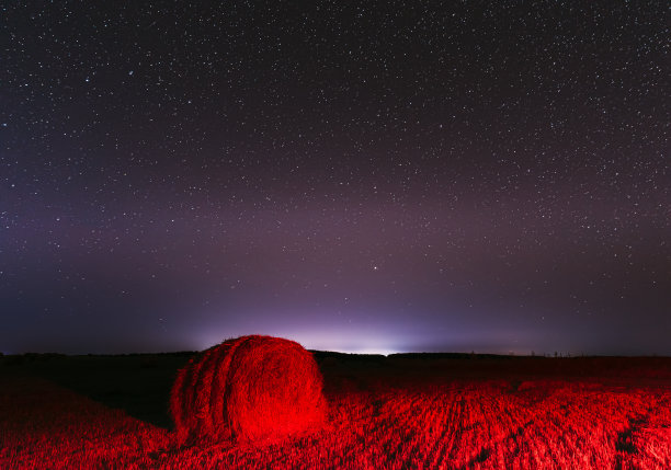 发光的星星和日落的灯光收获后的干草捆。农业彩色背景复制空间。夜晚星空之上的乡村景观草地与卷草在田里收获后图片下载