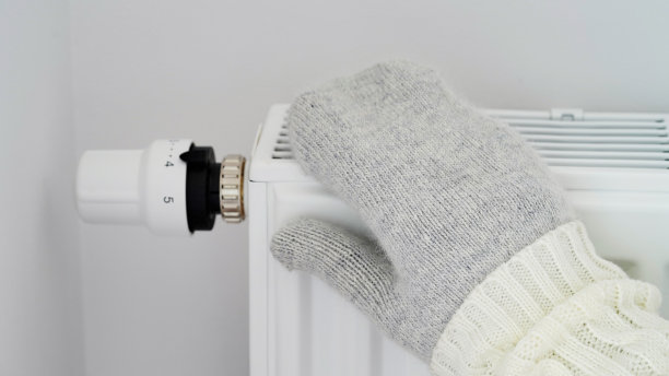 特写的女人在白色的毛衣和灰色的连指手套温暖她的手在暖气片对着白墙。微距照片女性的手，复制空间。冬季节气理念。欧洲的天然气危机。图片下载