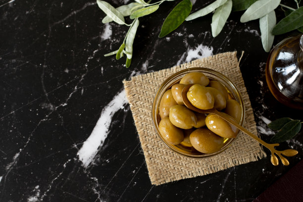 绿橄榄在一个玻璃碗和一个金勺子，特级初榨橄榄油，新鲜的树枝在黑暗的背景与复制空间图片下载