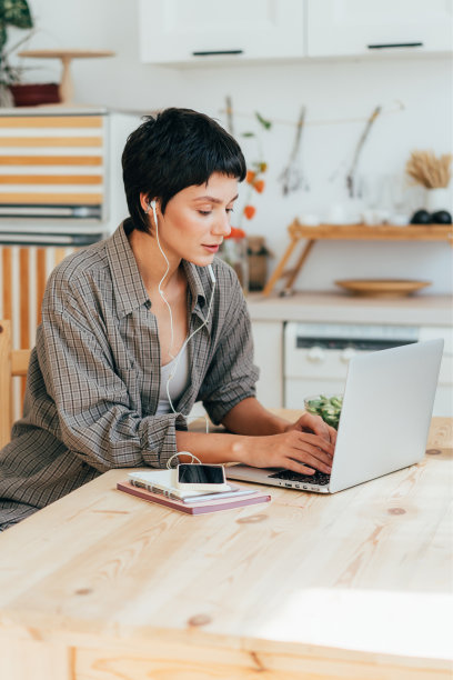 女孩在厨房用笔记本电脑工作，戴着耳机听音乐。图片下载