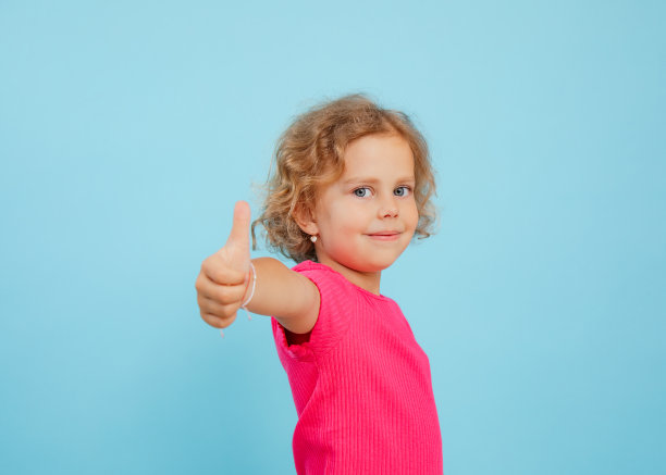 肖像微笑的小女孩，金发穿着粉红色的t恤，看着，显示大拇指在蓝色背景上的标志。图片下载