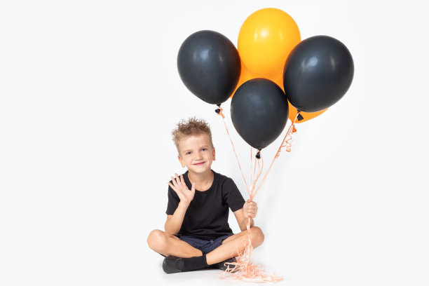 一个戴着巫师帽的快乐男孩在迎接一只手，手里拿着黑色和橙色的气球，坐在白色的画室背景上图片下载