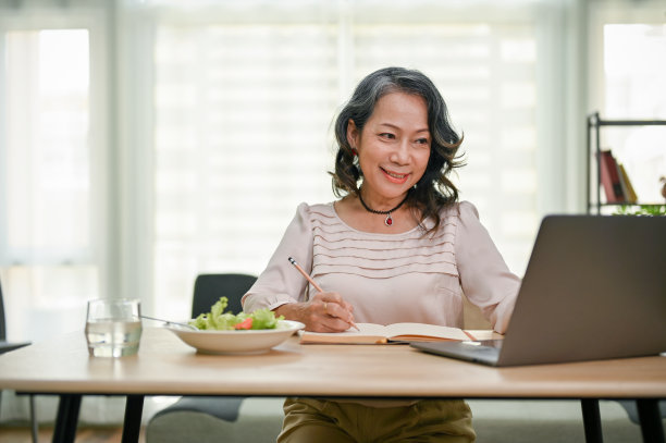 快乐的亚裔女商人在书上写着什么，看着笔记本电脑屏幕。图片下载