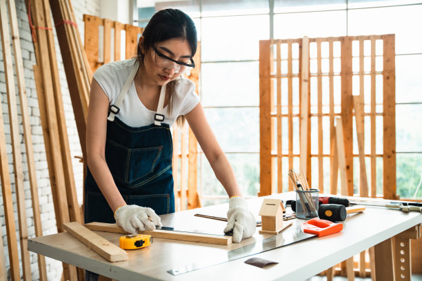 戴着手套和护目镜的女木匠测量木材的比例，为制作产品准备零件。图片下载