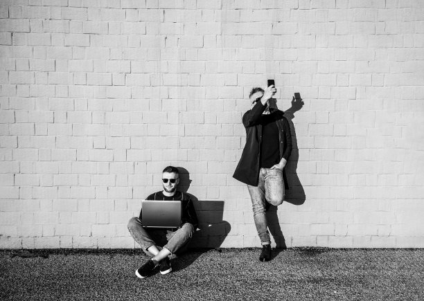 两个朋友在午休时间拿着数码设备——年轻的白人男子盘腿坐在地上，用笔记本电脑工作——男子站着用智能手机自拍——黑白滤镜图片下载