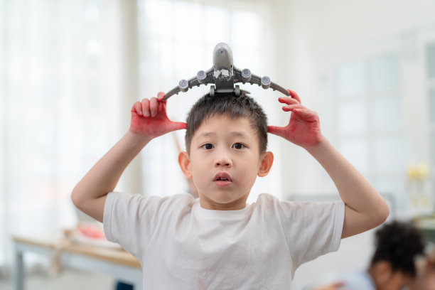 在国际学校的教室里，年轻的亚洲男孩把飞机模型戴在头上。看相机。在课堂上玩耍感觉快乐和享受。回到学校和教育理念图片下载