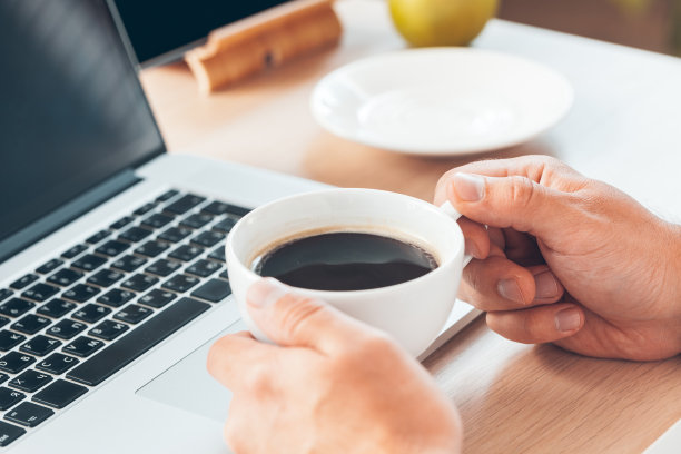 年轻的商人拿着热咖啡在笔记本电脑前工作。图片下载