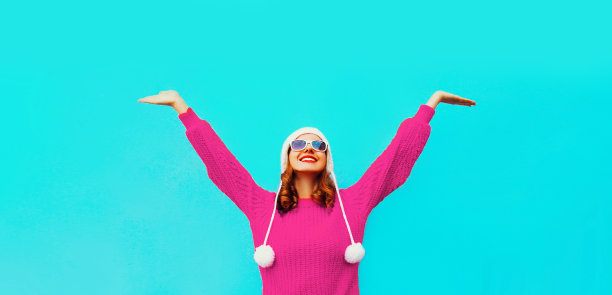 快乐的微笑着的妇女举起她的手，穿着冬天的粉红色针织毛衣，帽子上有蓝色背景的棒棒糖图片下载