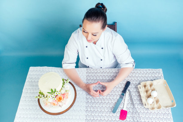 糕点师女厨师展示手指心形符号旁边的奶油白色两层婚礼(生日)蛋糕与鲜花在工作室的桌子上蓝色背景。热爱工作的概念图片下载