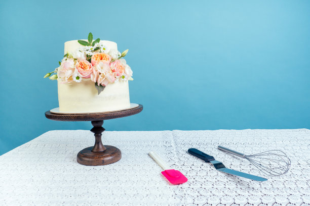 Copyspace模型工作空间面包师糕点厨师奶油白色双层婚礼(生日)蛋糕与鲜花，工具和打蛋器在桌子上和工作室的饼干蓝色背景图片下载