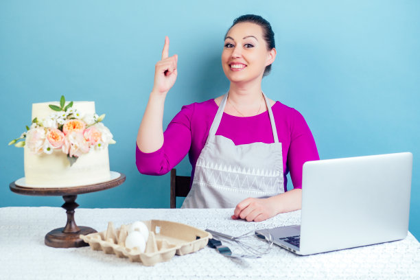 糕点家庭主妇(糕点厨师)的女人指着旁边的奶油白色两层婚礼蛋糕，鲜花和笔记本电脑在工作室的桌子上，蓝色的背景。图片下载