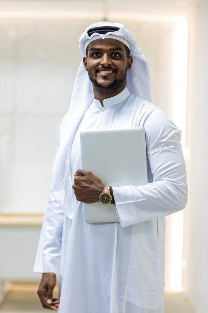 身着传统白色坎杜拉服装的穆斯林商人站在他现代化的办公室里，手里拿着笔记本电脑图片下载