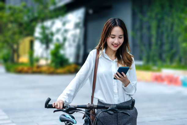 年轻的亚洲女商人骑着自行车，用手机站在城市街道上，自行车看着手机。图片下载