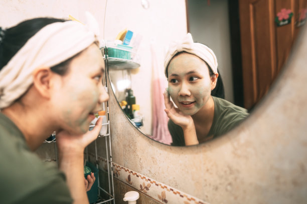 年轻的亚洲妇女清洁脸与绿色粘土面膜愈合痤疮反射与镜子在浴室图片下载