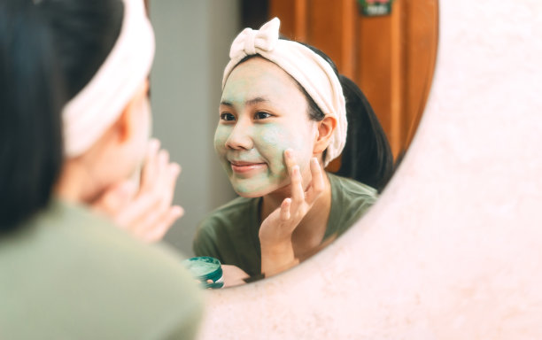 年轻的亚洲妇女清洁脸与绿色粘土面膜愈合痤疮反射与镜子在浴室图片下载