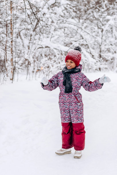 一个快乐的非裔美国小女孩，戴着红帽子，穿着连体衣，手里拿着一个燃烧的火花，在冬天的森林里。冬天的乐趣，圣诞快乐和新年快乐的概念图片下载