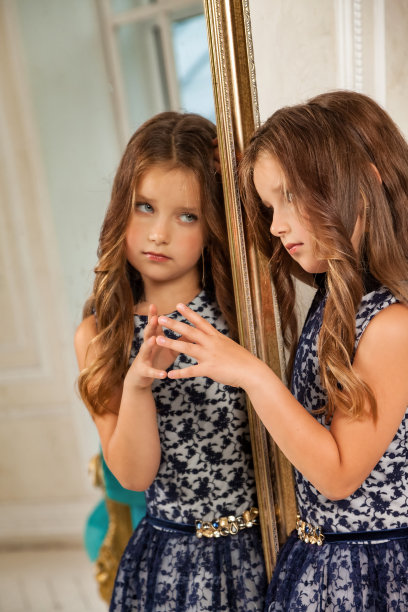 画像小女孩公主5-6岁，思考，看着反射，时尚模特在时尚优雅的蓝色连衣裙在客厅与镜子。时尚年轻女演员在家里。本空间图片下载