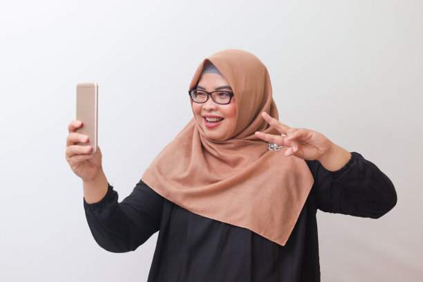 戴着头巾的亚洲妇女在智能手机上自拍，表情积极，手势和平图片下载