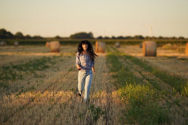 这个女孩在夏天跑过田野，黑色的长发在风中飘动，她穿着牛仔裤和浅色的衬衫，围着一堆干草堆和割过的草。自然农业图片下载