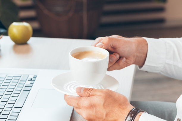 年轻的商人拿着热咖啡在笔记本电脑前工作。图片下载